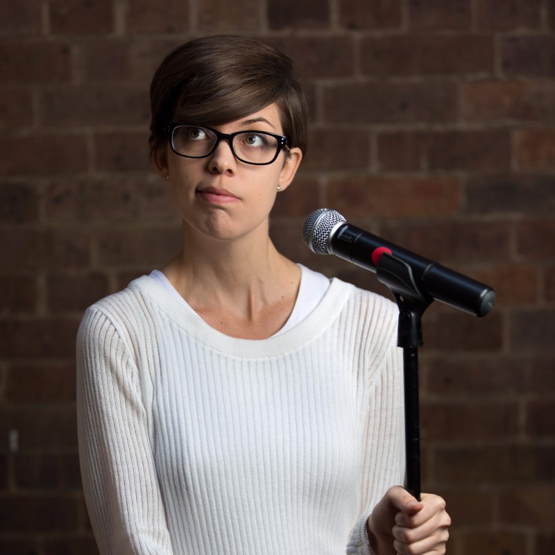 Kaitlyn holding a microphone stand and looking up with a questioning expression, standing in front of a red brick wall.
