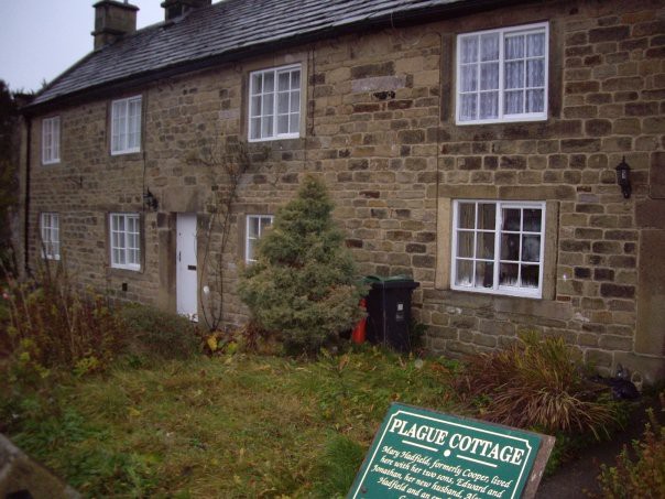 A two-storey stone cottage with white square windows and grassy front yard. A large green plaque with white writing on it says “PLAGUE COTTAGE”.