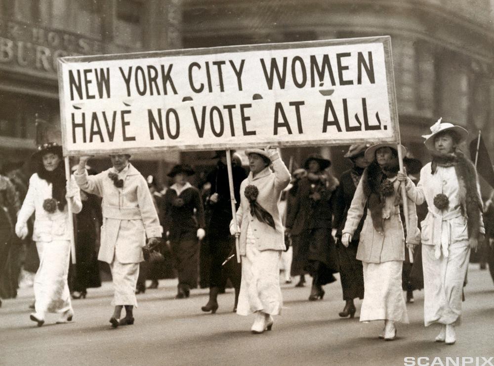 Black-and-white photograph of five women wearing white Victorian-era dresses, hats and rosettes, walking in a protest march while holding a banner that says “NEW YORK CITY WOMEN HAVE NO VOTE AT ALL”.